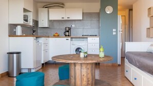 Eine kompakte Küche mit Mikrowelle, Kaffeemaschine und Spüle. Ein runder Holztisch mit Pflanze und Flasche steht dort. Blaue Sitzsäcke liegen auf dem Boden.