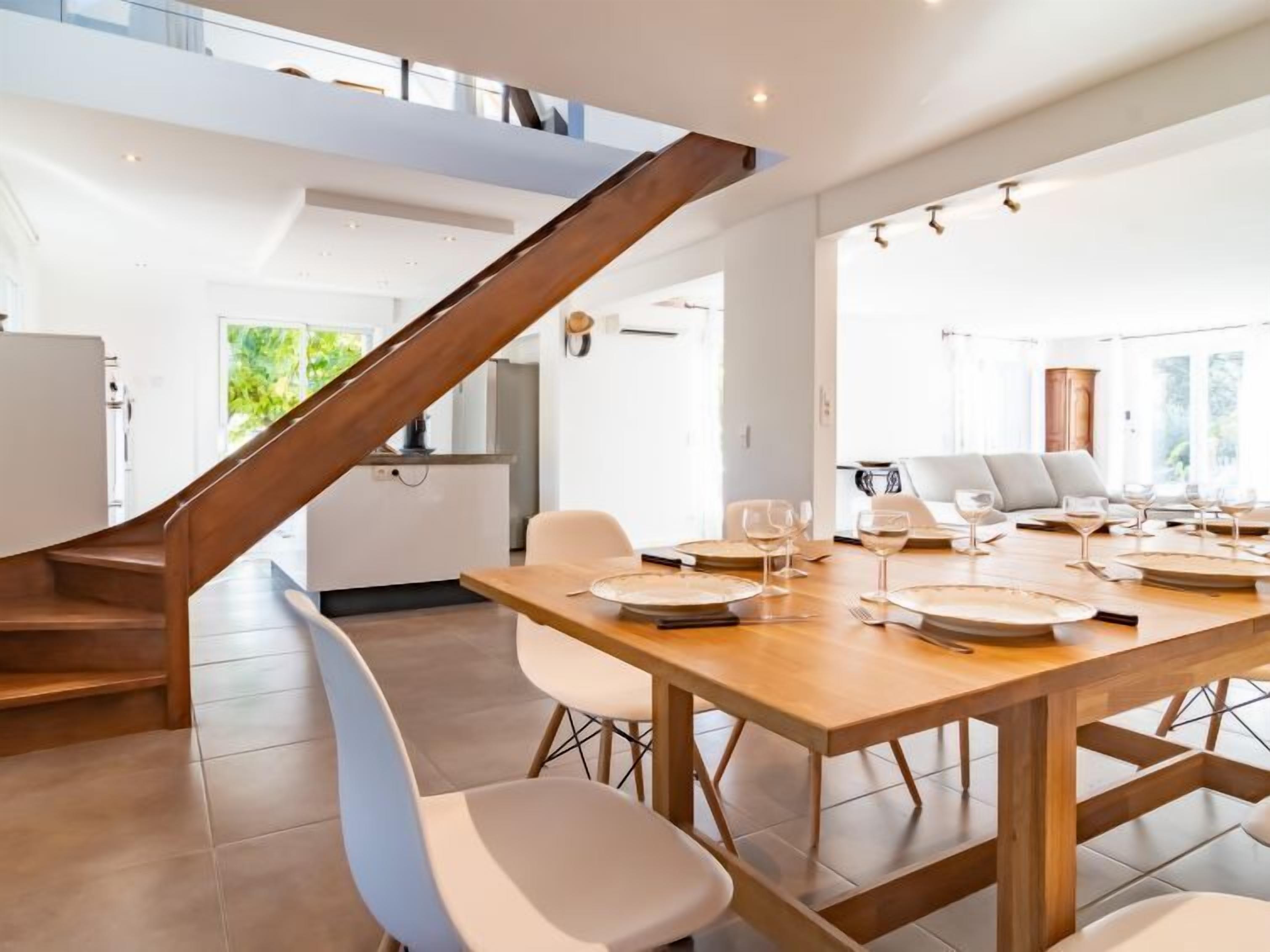 A modern kitchen with a wooden staircase, a dining area with a large wooden table set for six, and a view of the outdoors.