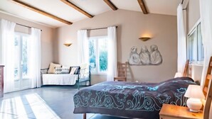 Un dormitorio amplio con una cama grande, una mesita de noche de madera y una lámpara de pared. Se aprecia una ventana con cortinas, una silla y un cuadro en la pared.