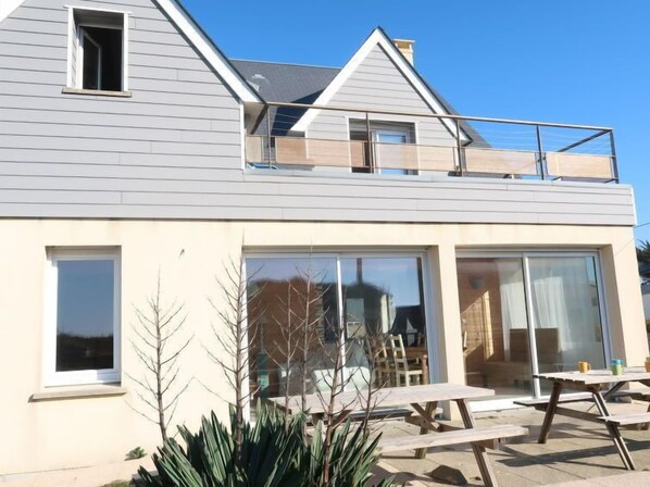 Une maison moderne à deux étages, dotée d’un balcon, de grandes portes-fenêtres coulissantes et d’un patio aménagé avec des tables de pique-nique.