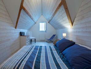 Une chambre avec un plafond en pente, un lit avec une literie à rayures, une chaise, un bureau avec une lampe et une fenêtre.