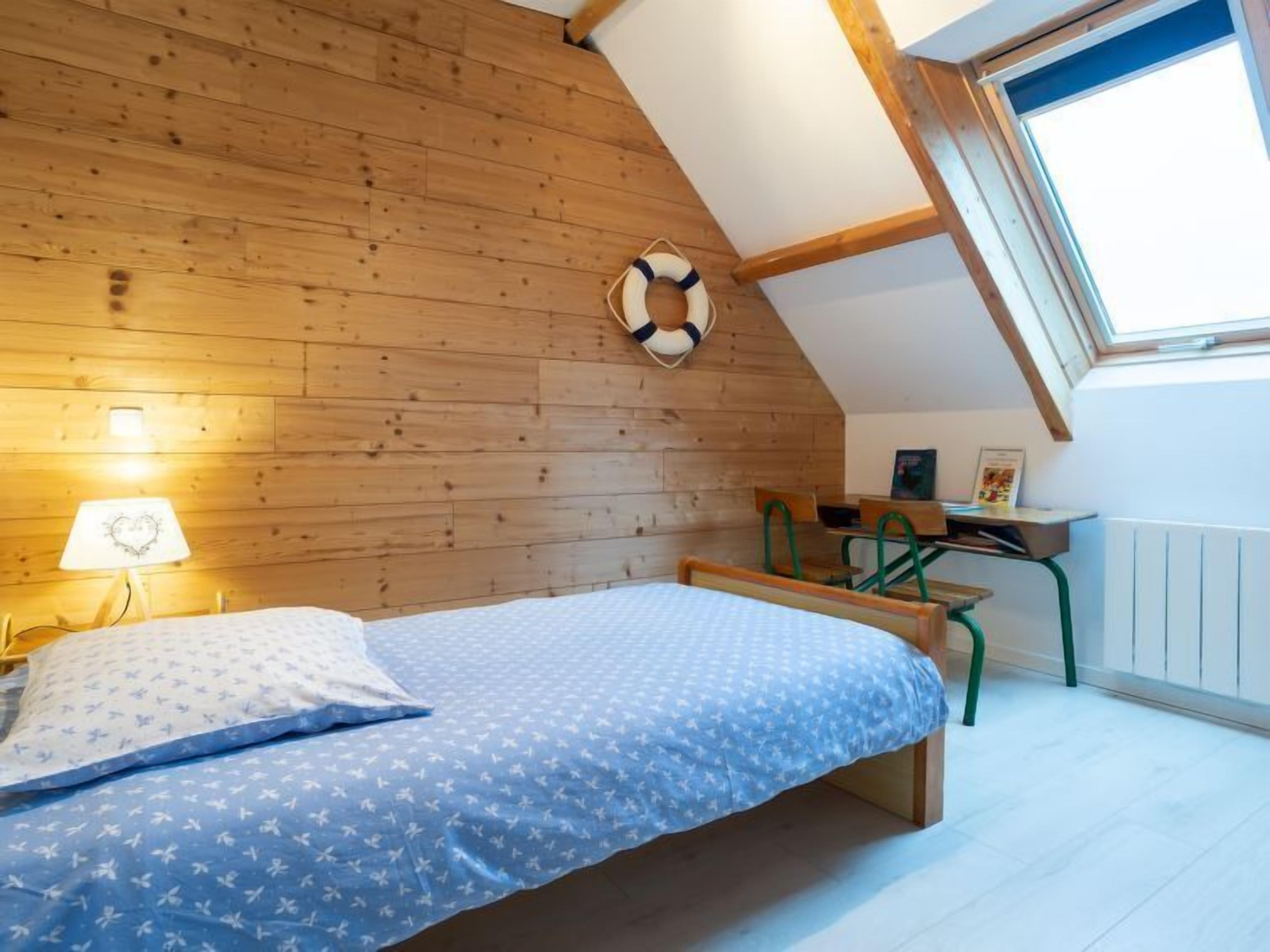 Ein gemütliches Schlafzimmer mit schrägen Wänden, Holzvertäfelung, einem Bett mit blumigem, blauem Überwurf, einem Schreibtisch mit Stuhl und einem Fenster mit Blick.