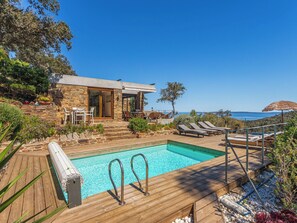 A poolside area with a wooden deck, lounge chairs, and a stone building with a glass door.