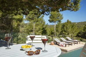 A poolside table with wine glasses, a bowl of fruit, and sunglasses. There are lounge chairs and a pool in the background.