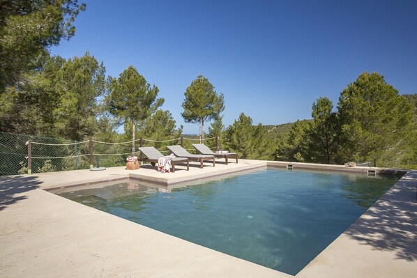 A rectangular swimming pool with clear blue water, surrounded by a concrete deck. There are three lounge chairs and a small wooden bench. The pool area is enclosed by a chain-link fence, and trees are visible in the background.