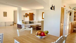 Ein geräumiges Wohnzimmer mit einem weißen Sofa, einem Holztisch mit Obstschale und einer Küchenzeile mit Holzschränken und einem weißen Kühlschrank.