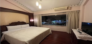 Ein Hotelzimmer mit einem großen Bett, einem Fernseher und einem Fenster mit Blick auf die beleuchtete Stadt.