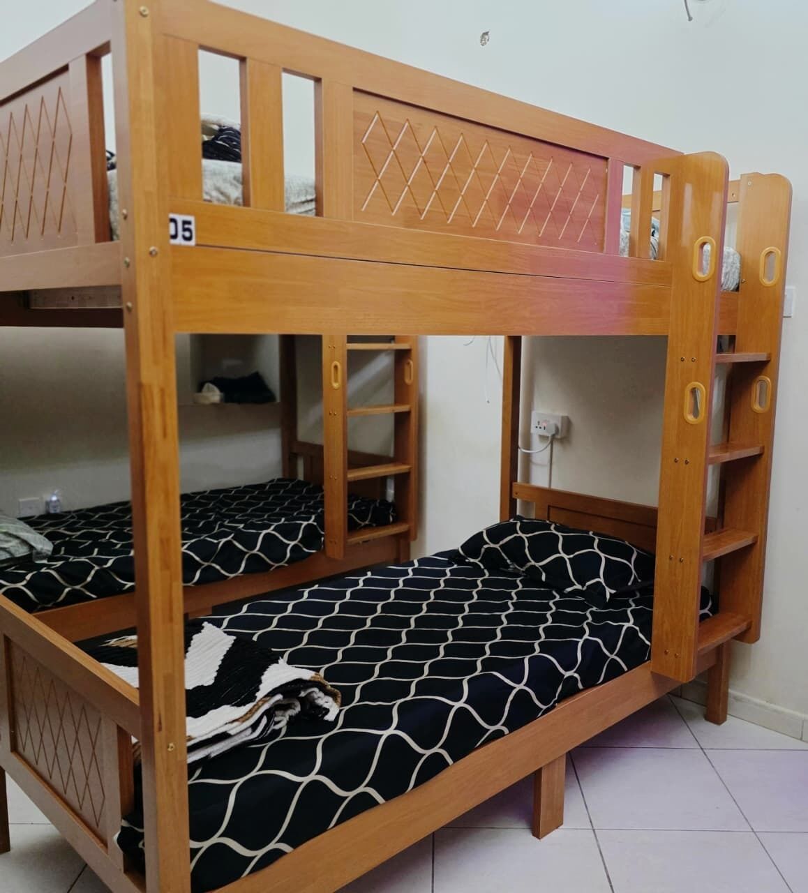 A bunk bed with a wooden frame and a patterned mattress. There are shelves built into the bed frame, and a power outlet is visible on the wall.