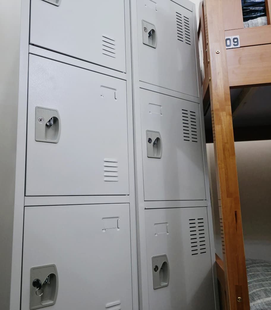 Een aantal witte metalen kasten met ventilatieopeningen, een houten stapelbed met het nummer 09, en een gedeeltelijk zicht op een bed met een bedekking met patroon.