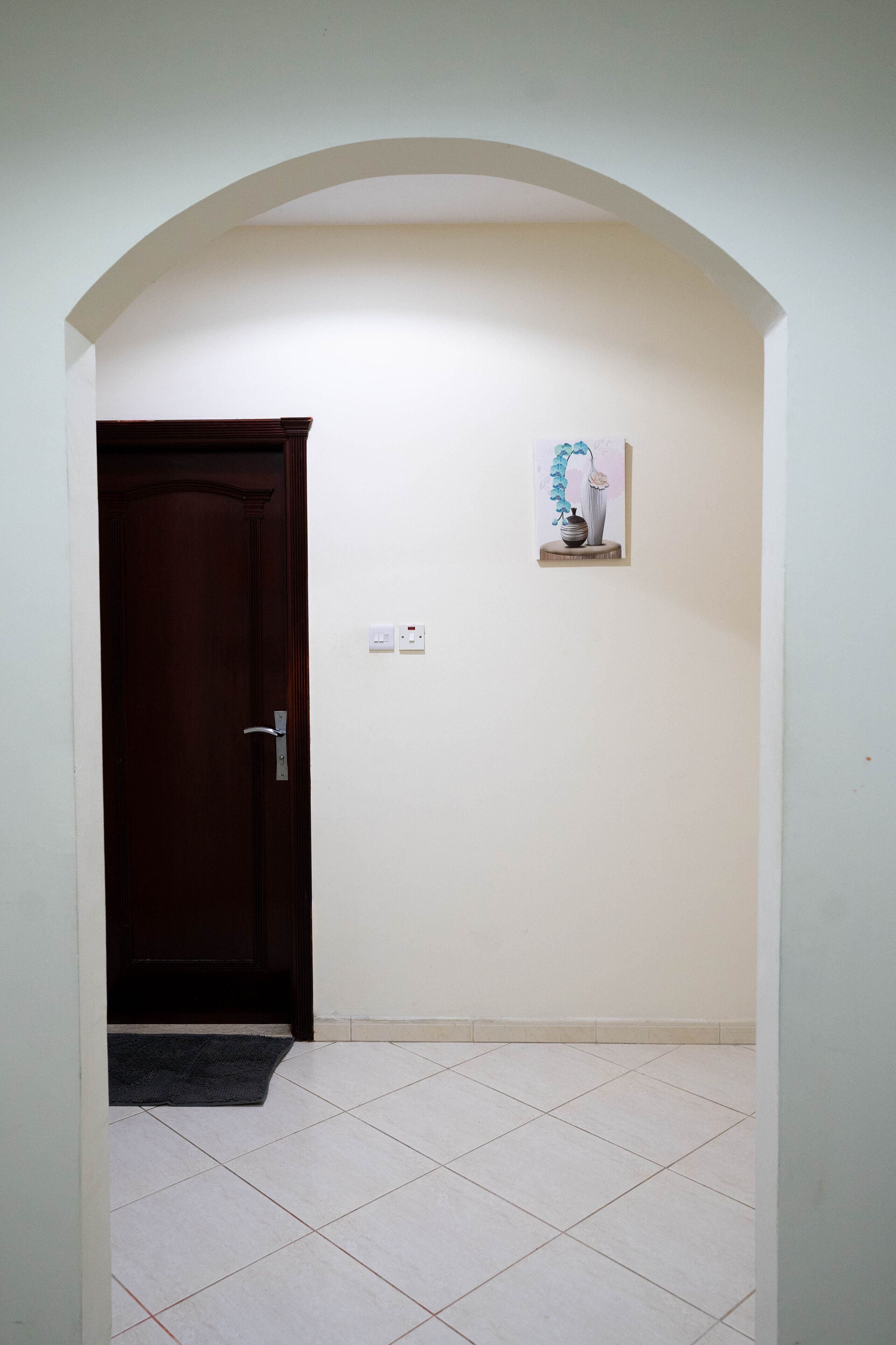Um corredor com porta de madeira, uma pequena estante com um vaso e chão de azulejos.