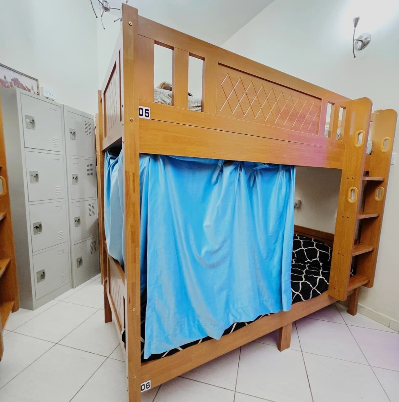 Ein Stockbett mit blauer Vorhang, Holz-Kopfteil und einer Stehle. Es gibt Schließfächer und einen Fliesenboden.