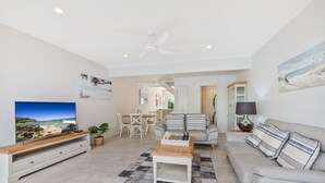 A spacious living room with a flat-screen TV, a wooden coffee table, a grey sofa, and a dining area with white furniture. - Unit 37 - 2 Bedroom Deluxe Waterfront Townhouse (Noosaville)