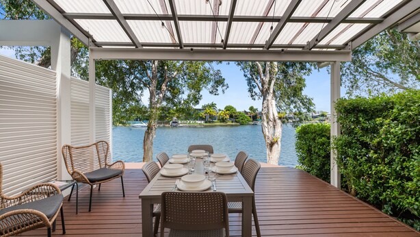 A covered outdoor dining area with a table set for six, overlooking a lake with trees and a boat in the background. - Unit 37 - 2 Bedroom Deluxe Waterfront Townhouse (Noosaville)