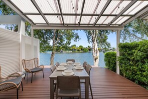 A covered outdoor dining area with a table set for six, overlooking a lake with trees and a boat in the background.