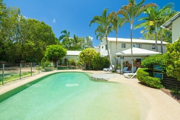 A swimming pool with a curved edge, surrounded by palm trees and a building with a white roof.