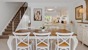 Un comedor con mesa de madera y sillas blancas, una escalera y una cocina al fondo.