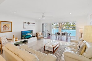 A modern living room with a television, comfortable seating, a glass coffee table, and a view of a pool and outdoor area. - Unit 31 - 3 Bedroom Deluxe Waterfront Townhouse (Noosa)