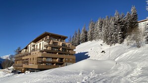 Un hotel de varios pisos con balcones, ubicado en una pendiente nevada bajo un cielo azul despejado.