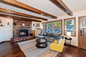 A living room with a brick fireplace, a sectional sofa, a yellow armchair, and large windows. - Bufflehead Cottage by Destination Big Bear: Pool Table! Spa! Mountain Views! (Big Bear City)