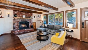 A spacious living room with a brick fireplace, wooden beams, a gray sofa, a yellow armchair, and large windows offering a view of the outdoors.