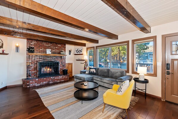 Amplia sala de estar con chimenea de ladrillo, vigas de madera, un sofá gris, un sillón amarillo y ventanas grandes con vistas al exterior.