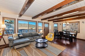 A spacious living room with a gray sofa, a yellow armchair, a dining table with chairs, and large windows offering a view of the outdoors. - Bufflehead Cottage by Destination Big Bear: Pool Table! Spa! Mountain Views! (Big Bear City)