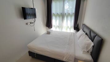 A neatly made bed with white linens, a flat-screen TV mounted on the wall, and a window with sheer curtains.