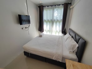 A neatly made bed with white linens, a flat-screen TV mounted on the wall, and a window with sheer curtains.