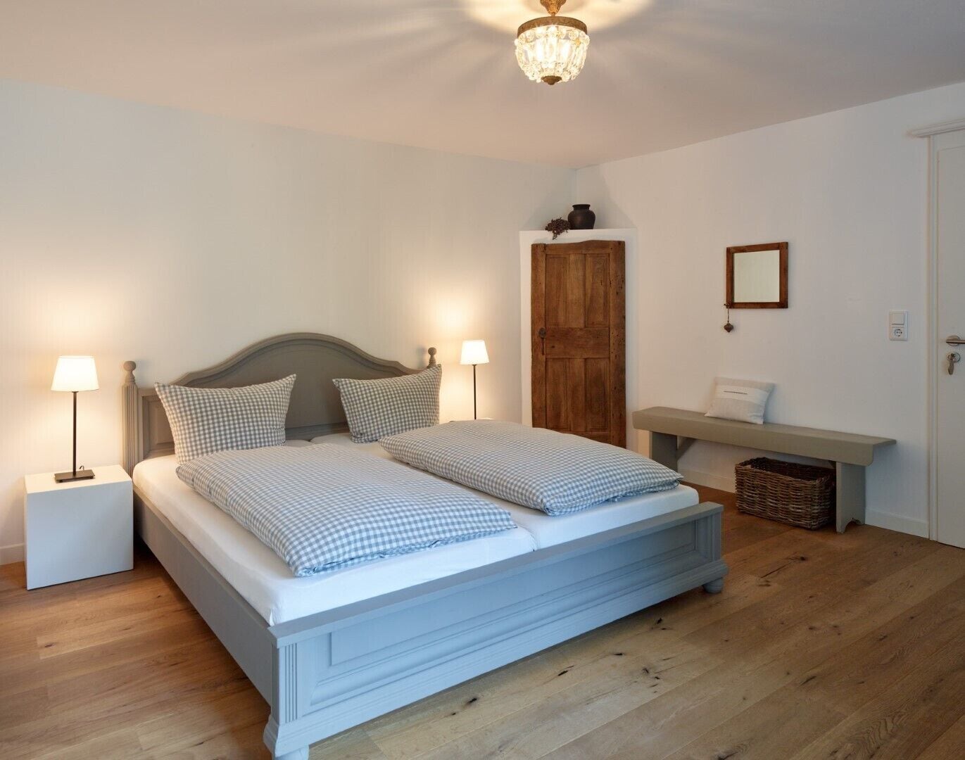 Ein Doppelbett mit karierter Bettwäsche, zwei Nachttischlampen, eine Holztür, eine Bank und ein kleiner Korb auf dem Boden.