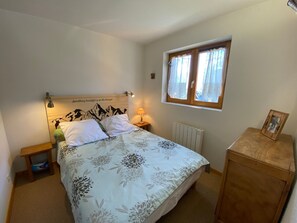 A bedroom with a bed, a nightstand, a bedside lamp, a window with curtains, and a radiator. - Cozy apartment with terrace, near village, parking included (Châtel)