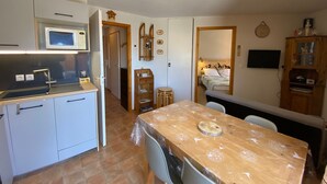Une cuisine compacte équipée d’un four à micro-ondes, d’un évier et de placards, une table à manger avec des chaises, un petit tabouret, un lit avec une couvre-lit à motifs de montagne, une télévision et un meuble en bois contenant des objets décoratifs.