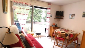 Una sala de estar con un sofá rojo, una mesa de comedor con sillas, un televisor montado en la pared y una ventana con cortinas.