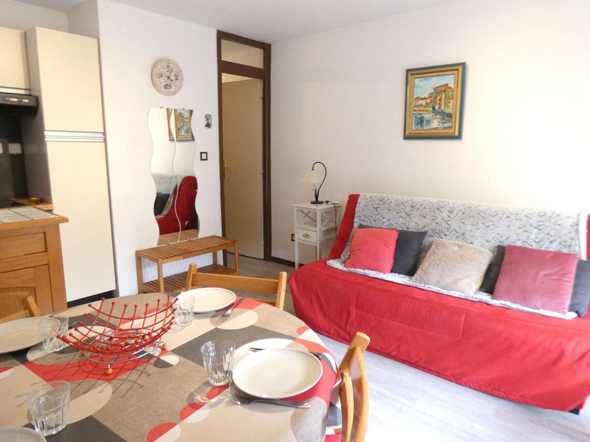 Una habitación con una mesa de comedor para dos personas, un sofá con cojines rojos y grises, una mesita de noche blanca y un cuadro en la pared.