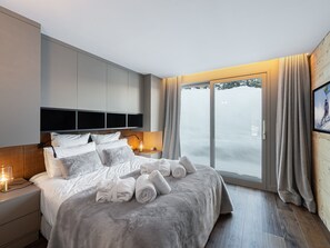 Ein modernes Hotelzimmer mit einem großen Bett, einem Nachttisch mit Lampe, einem Kleiderschrank und Blick auf eine verschneite Landschaft durch eine Glastür.