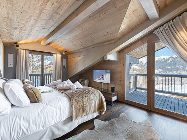 A bedroom with a large bed, a television, and a view of mountains through a balcony.