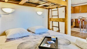 Een slaapkamer met een hoogslaper, houten balken, twee bedlampen, een dienblad met foto’s en uitzicht op de keuken.