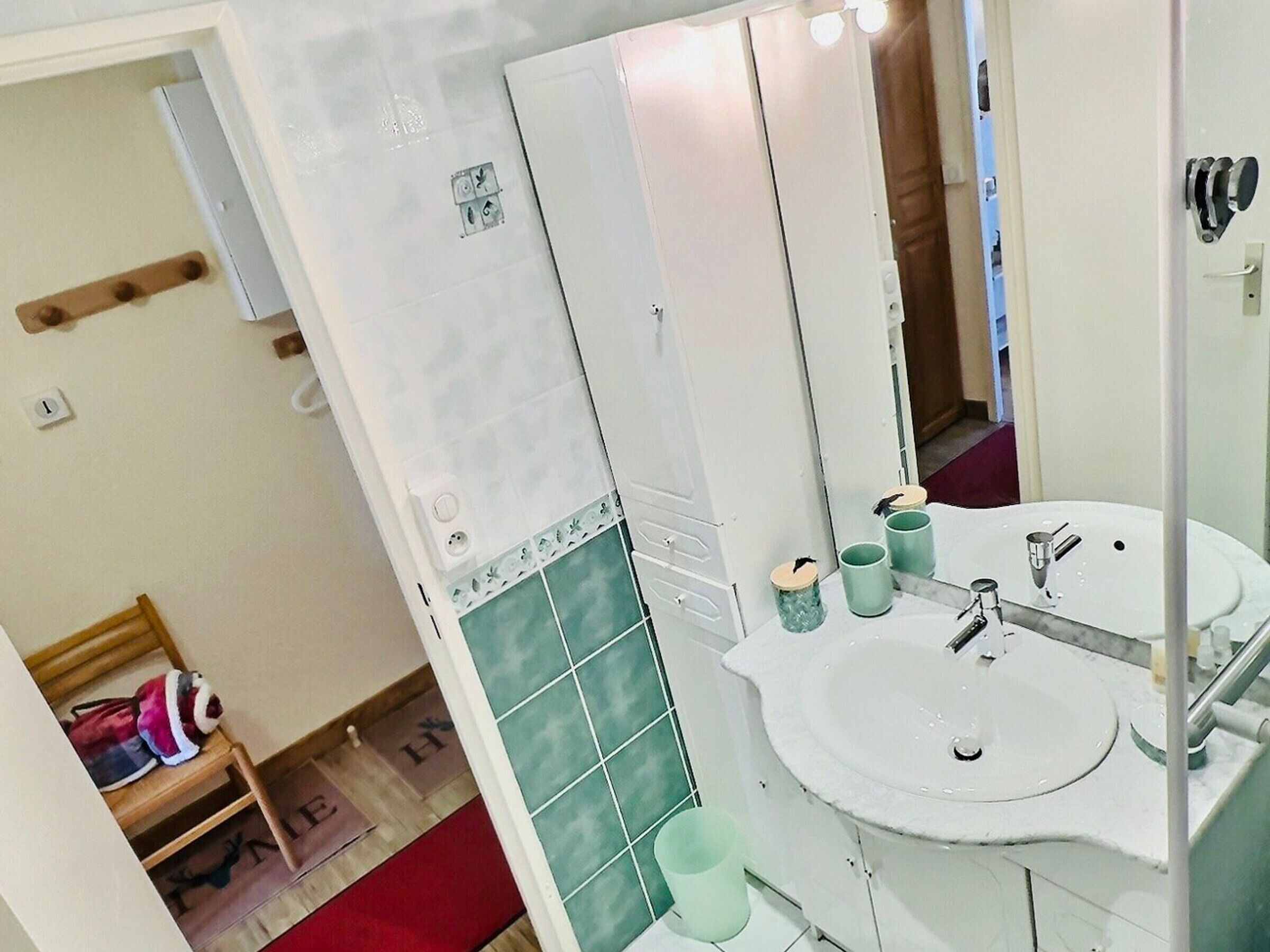 Une salle de bain avec un lavabo blanc, un miroir et un mur recouvert de carreaux verts. On y trouve une étagère en bois sur laquelle est posé un sac, ainsi qu’un tapis rouge sur le sol.