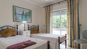 Ein Schlafzimmer mit zwei Betten, einem Nachttisch, einem Sessel und einem Fenster mit Vorhängen.