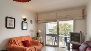 Ein Wohnzimmer mit einer Couch, einem Fernseher und einem Tisch mit Lampe. An der Wand befinden sich Bilder in Rahmen, und es gibt einen Balkon mit Tisch und Stühlen.