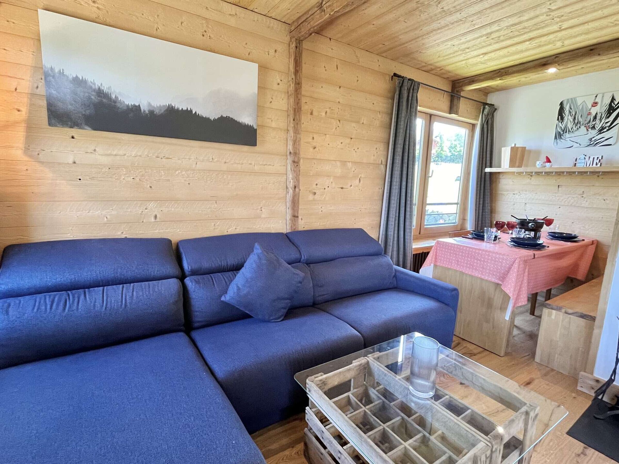 A cozy room with a blue sofa, a glass coffee table, wooden walls, and a dining area with a pink tablecloth.