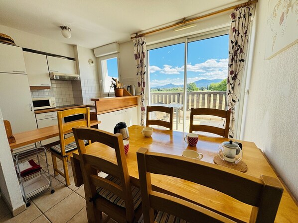 A dining area with a table and chairs, a view of the outdoors, a kitchen area with cabinets and a microwave, and a ceiling light fixture.