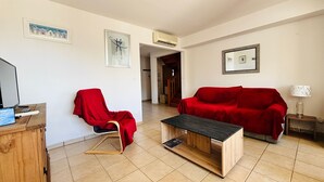 A living room with a red chair, a red sofa, a wooden coffee table, and a TV on a wooden cabinet. - Saint Cyprien - 2-Bedroom Apartment for 6 - Shared Pool, Terrace, Parking (Saint-Cyprien)