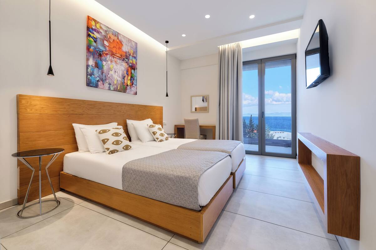Ein modernes Hotelzimmer mit einem großen Bett, einem Nachttisch, einem Gemälde an der Wand und Meerblick durch das Fenster.