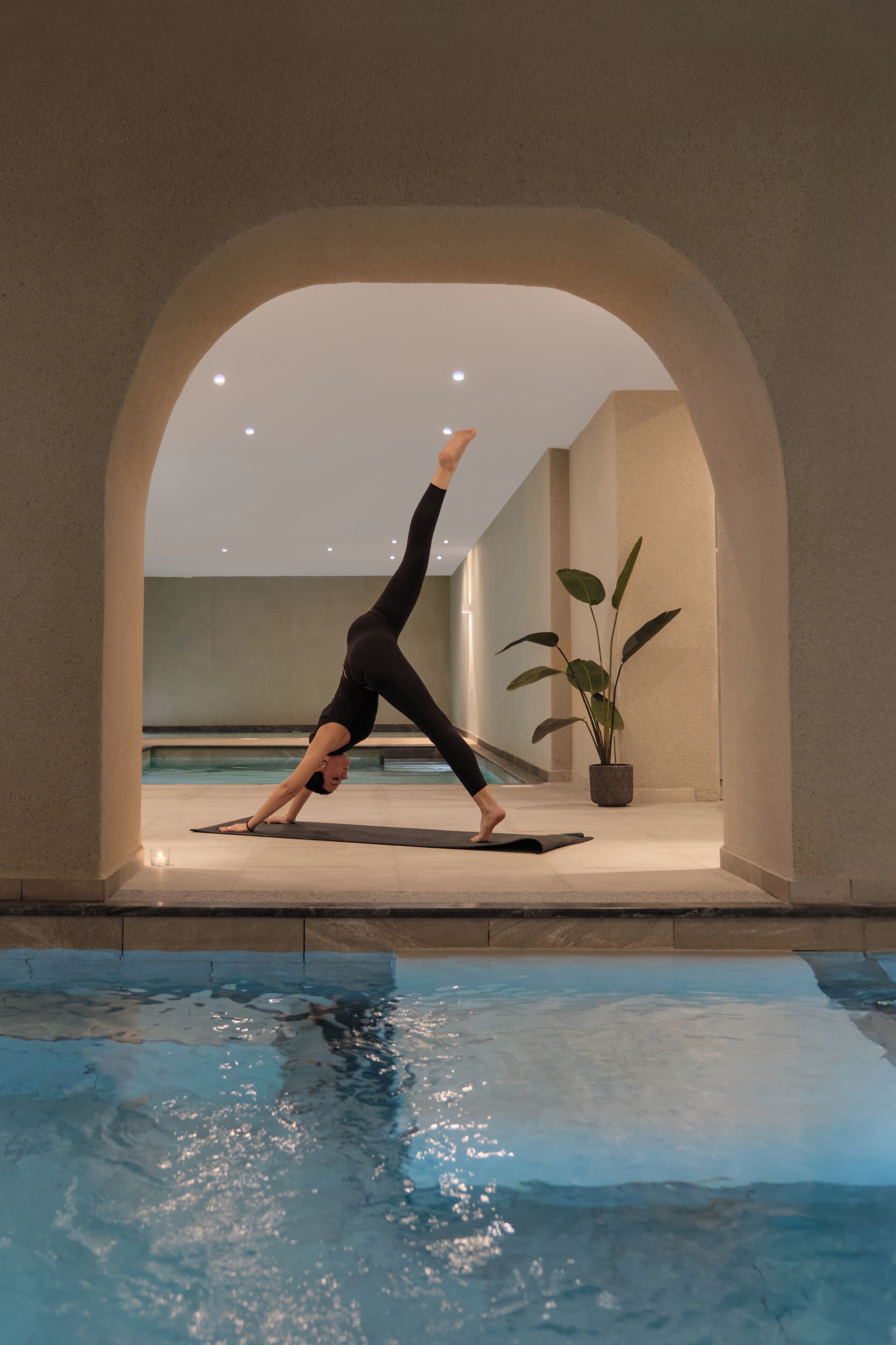 Una persona realizando una postura de yoga en un pasillo con un arco que da a una zona de piscina.