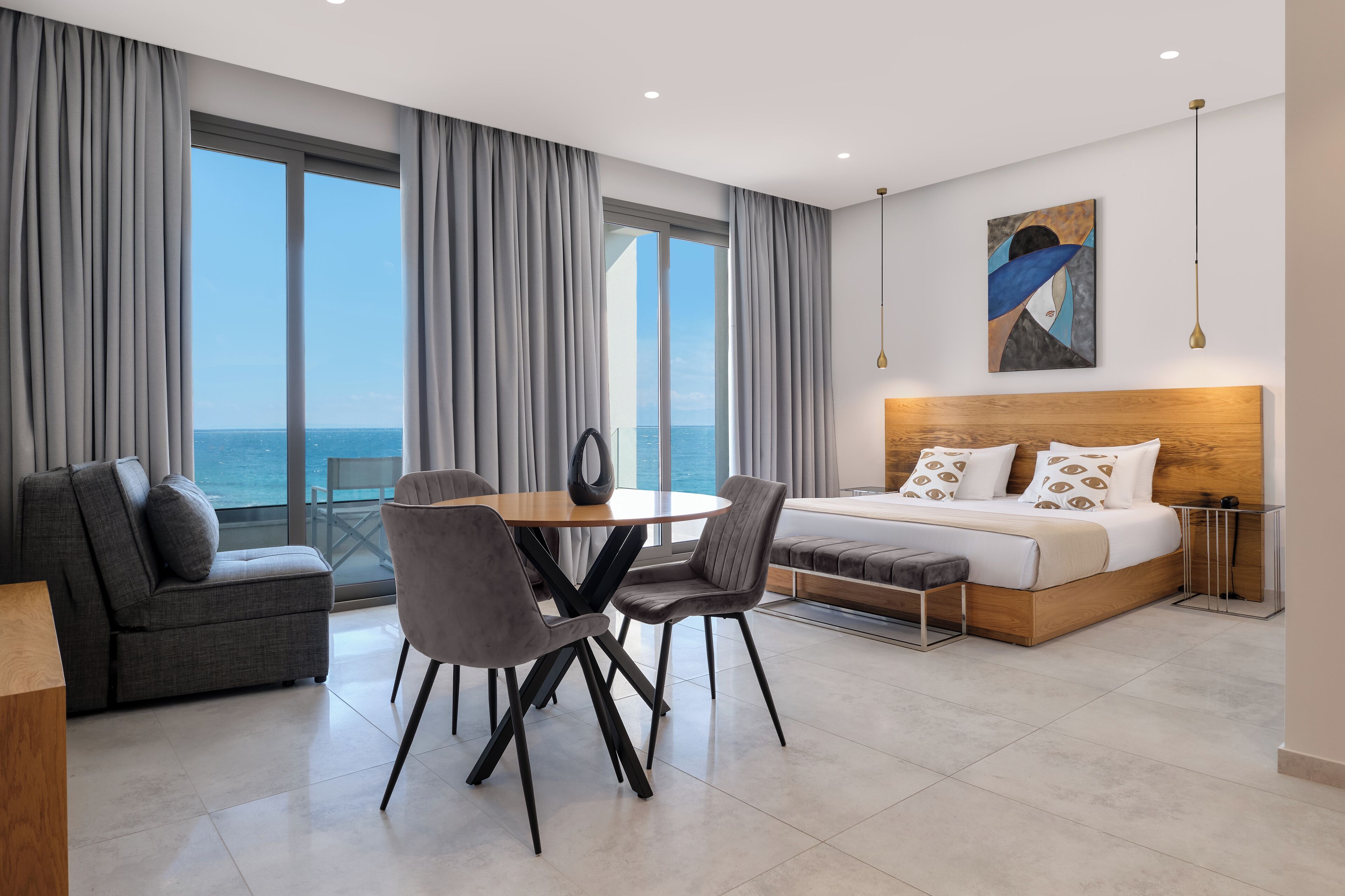 Una habitación de hotel moderna con una cama grande, un rincón de comedor con una mesa redonda y sillas, un sofá, y vistas al mar a través de amplias ventanas.