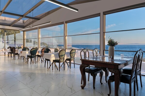 A dining area with tables and chairs, large windows with a view of the sea, and a modern ceiling design. - Mythical Coast Wellness Retreat (Lesvos)