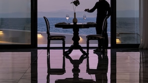 A dining area with a table and chairs, a vase with a flower, and a view of the sea through large windows. - Mythical Coast Wellness Retreat (Lesvos)