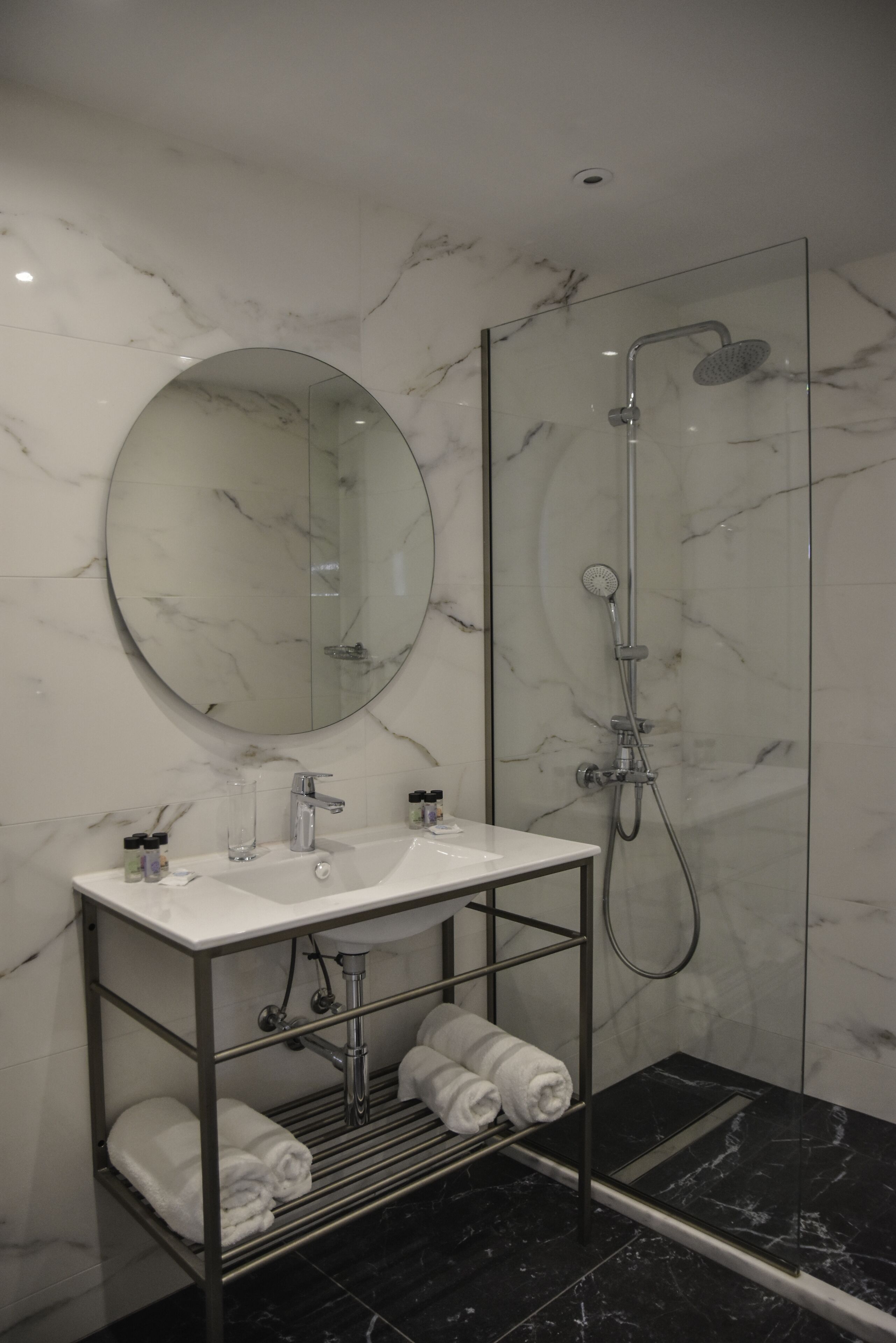 Un baño moderno con tocador de mármol, un espejo redondo, cabina de ducha con paredes de vidrio y toallas ordenadas en un perchero.