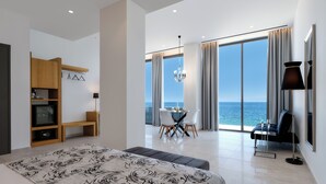 A modern hotel room with a large bed, a dining area with a view of the sea, and a balcony with a table and chairs. - Mythical Coast Wellness Retreat (Lesvos)