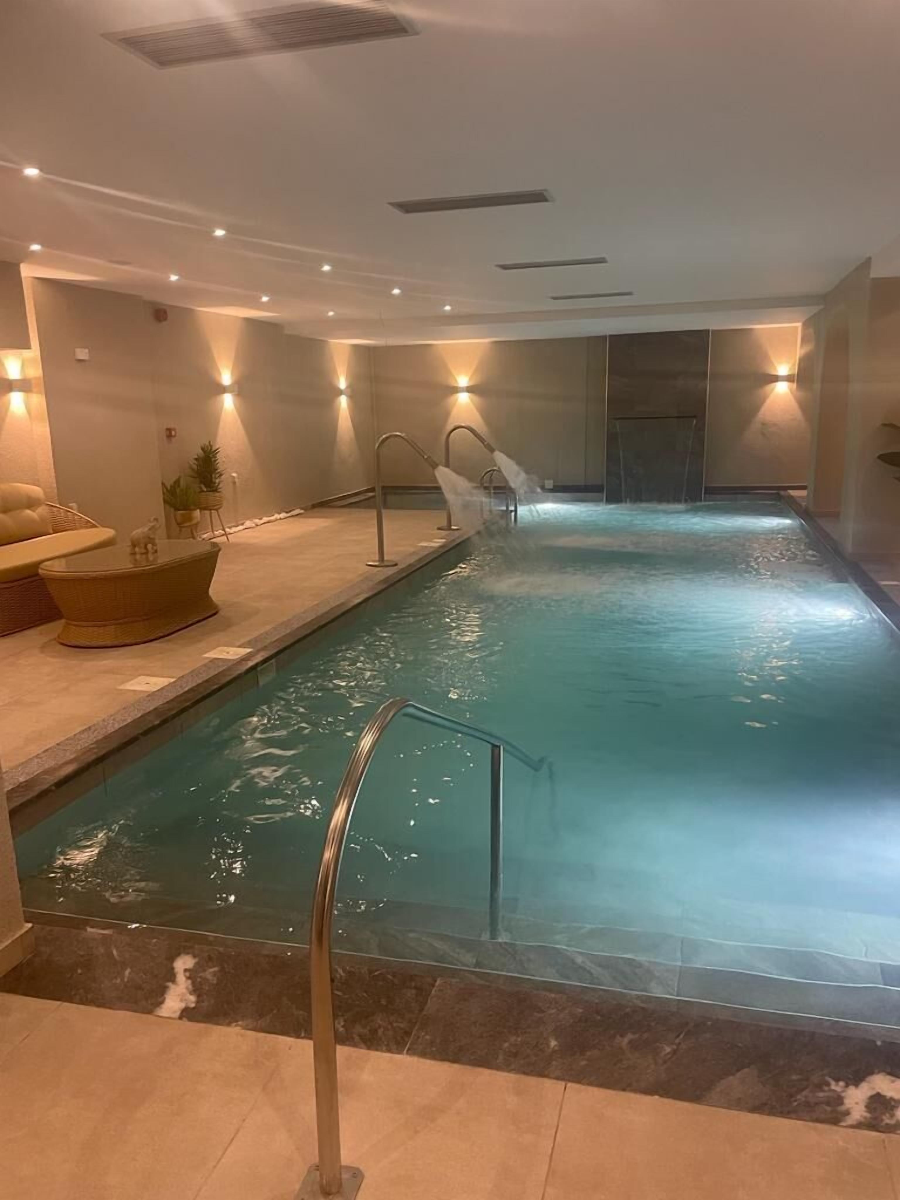 A swimming pool with a glass barrier, a lounge area with a sofa and a small table, and wall-mounted lighting.